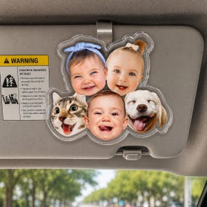 Puede incluir: Un accesorio de acr&iacute;lico transparente para el parasol del coche que presenta un collage de cinco retratos circulares, incluyendo tres beb&eacute;s y dos animales. Los beb&eacute;s tienen diversas expresiones y accesorios como una diadema azul y un lazo blanco.