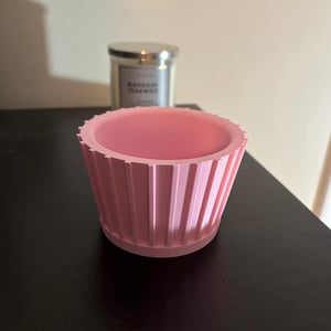 Peut inclure: Un pot de fleur rose clair, côtelé, avec une soucoupe assortie. Le pot a une forme cylindrique avec des rainures verticales. Une bougie dans un pot en verre avec un couvercle argenté est à l'arrière-plan.