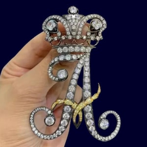 Peut inclure: Broche ornée en forme de lettre "A" surmontée d'une couronne, incrustée de diamants et de pierres jaunes. La broche est sertie dans un métal argenté. Le design est très détaillé et d'une esthétique royale.