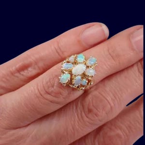 May include: A gold ring with a cluster of opals. The opals are oval-shaped and in shades of white and pale blue. The ring has a gold filigree design. The ring is shown on a person's finger.