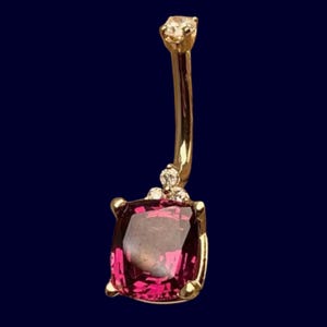 May include: A gold-coloured belly button ring featuring a large, square, magenta gemstone at the base. The ring has a curved design with a small, clear gemstone at the top and three small clear gemstones above the large gemstone.