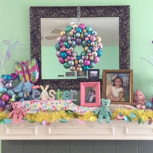 May include: A decorated mantelpiece with Easter-themed decorations. A wreath of colorful ornaments hangs above a display of stuffed animals, decorative eggs, and a sign that says "Easter". A mirror is in the background.