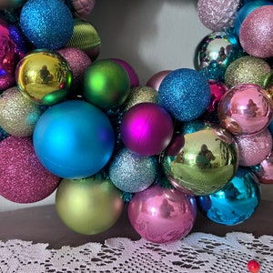 May include: A colorful Christmas wreath composed of various sized ornaments in shades of pink, blue, green, and silver. The ornaments have matte, glitter, and reflective finishes. The wreath is a festive holiday decoration.