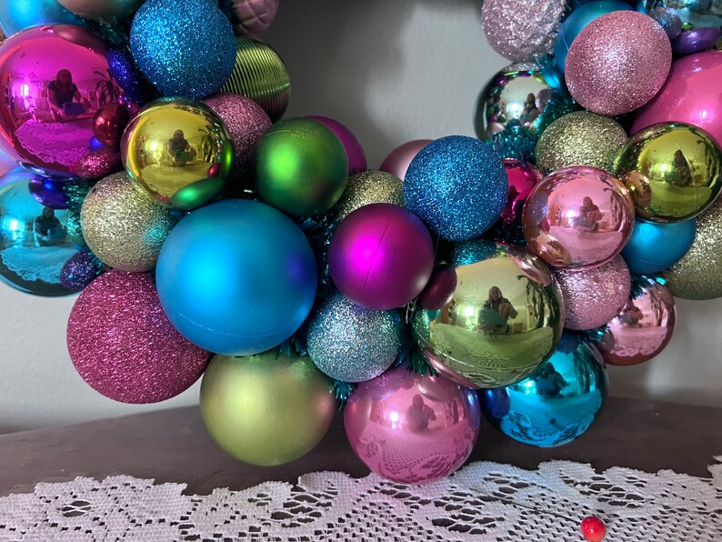 May include: A colorful Christmas wreath composed of various sized ornaments in shades of pink, blue, green, and silver. The ornaments have matte, glitter, and reflective finishes. The wreath is a festive holiday decoration.
