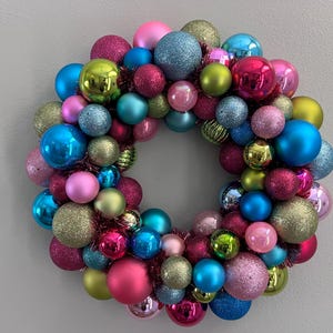 May include: A colorful Christmas wreath composed of various sized ornaments in shades of pink, blue, green, and gold. The ornaments have matte, glitter, and shiny finishes. The wreath is round and has a hole in the center.
