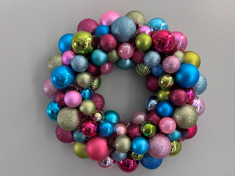 May include: A colorful Christmas wreath composed of various sized ornaments in shades of pink, blue, green, and gold. The ornaments have matte, glitter, and shiny finishes. The wreath is round and has a hole in the center.