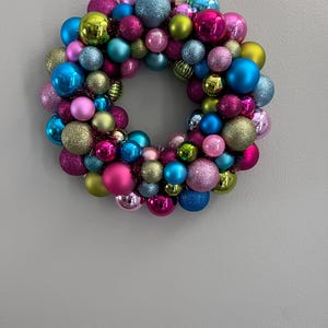 May include: A colorful Christmas wreath composed of various sized ornaments in shades of pink, blue, green, and gold. Some ornaments have glitter finishes, creating a festive and eye-catching holiday decoration.