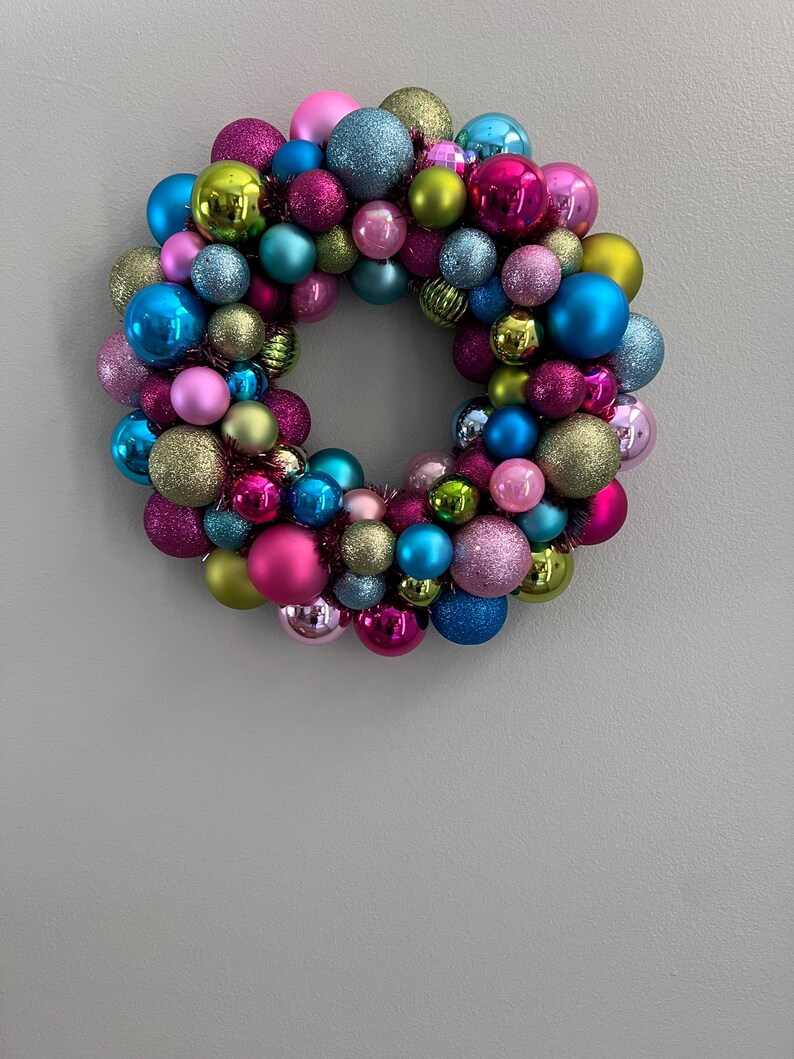 May include: A colorful Christmas wreath composed of various sized ornaments in shades of pink, blue, green, and gold. Some ornaments have glitter finishes, creating a festive and eye-catching holiday decoration.