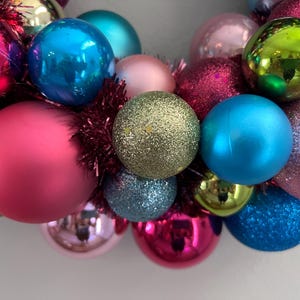 May include: A close-up of a colorful Christmas wreath. The wreath is composed of various sized ornaments in shades of pink, blue, green, and gold. Some ornaments are shiny, while others have glitter. Red tinsel fills the gaps.