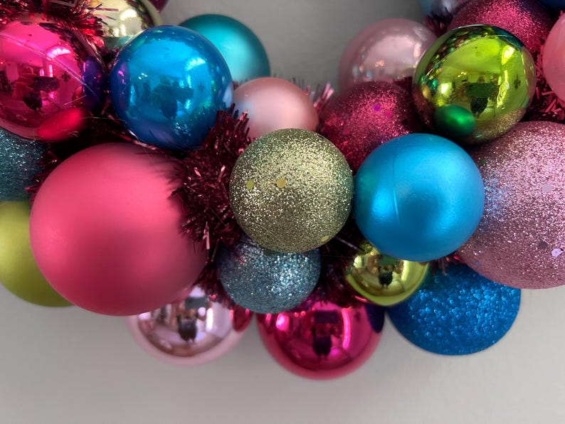 May include: A close-up of a colorful Christmas wreath. The wreath is composed of various sized ornaments in shades of pink, blue, green, and gold. Some ornaments are shiny, while others have glitter. Red tinsel fills the gaps.