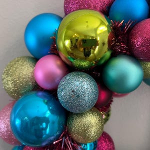 May include: A colorful Christmas wreath composed of various sized ornaments in shades of blue, pink, green, and gold. Some ornaments have a glittery finish, while others are smooth. The wreath is decorated with red tinsel.