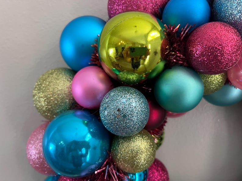 May include: A colorful Christmas wreath composed of various sized ornaments in shades of blue, pink, green, and gold. Some ornaments have a glittery finish, while others are smooth. The wreath is decorated with red tinsel.