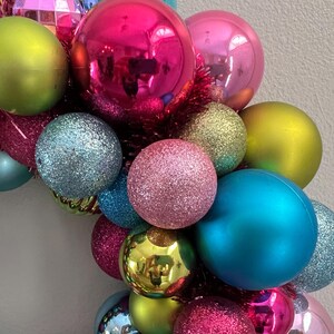 May include: A colorful Christmas wreath composed of various sized ornaments. The ornaments are in shades of pink, green, blue, and gold, with both matte and glitter finishes. The wreath is accented with red tinsel.