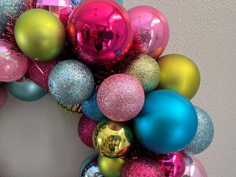 May include: A colorful Christmas wreath composed of various sized ornaments. The ornaments are in shades of pink, green, blue, and gold, with both matte and glitter finishes. The wreath is accented with red tinsel.