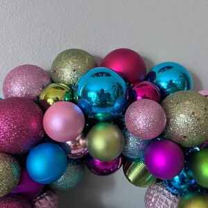 May include: A colorful Christmas ornament wreath composed of various sized spheres. The ornaments are in shades of pink, blue, green, and purple, with both matte and glitter finishes. The arrangement is festive and decorative.
