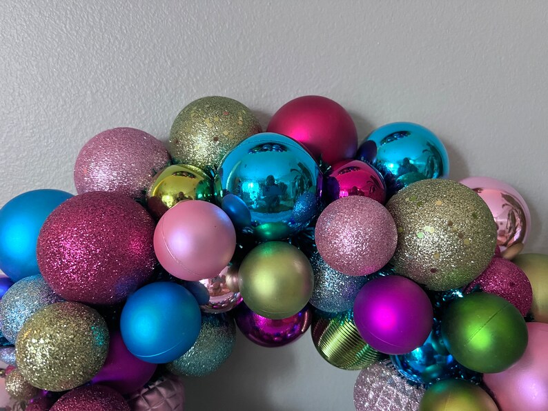 May include: A colorful Christmas ornament wreath composed of various sized spheres. The ornaments are in shades of pink, blue, green, and purple, with both matte and glitter finishes. The arrangement is festive and decorative.