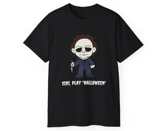 Może przedstawiać: Czarny t-shirt z kreskówkową ilustracją postaci trzymającej nóż. Postać ma białą maskę i nosi niebieski kombinezon. Tekst "SIRI, PLAY "HALLOWEEN"" jest wydrukowany poniżej.