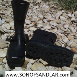 May include: Two black leather boots with buckles. The boots have a chunky sole with a tread pattern. The boots are on a background of small white rocks.