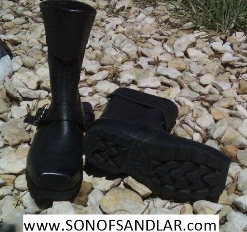 May include: Two black leather boots with buckles. The boots have a chunky sole with a tread pattern. The boots are on a background of small white rocks.