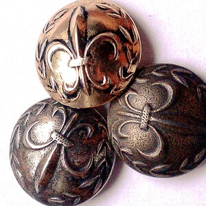 May include: Three antique metal buttons with a fleur-de-lis design. The buttons are in shades of gold and silver, with intricate detailing. The buttons are round and have a slightly domed shape, suggesting they are vintage or antique.