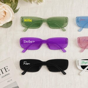 May include: Six pairs of rectangular sunglasses in green, blue, purple, pink, black, and white. The lenses and frames are translucent except for the solid white pair. Each pair features a custom name printed in a script or block font on the lens.