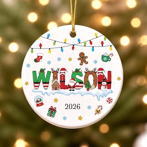May include: A round white ceramic Christmas ornament featuring the name WILSON in colorful, patterned letters. The design includes holiday icons like a gingerbread man, Santa, a snowman, and string lights. The year 2026 is printed in black.