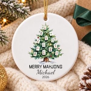 May include: A round white ceramic ornament featuring a green Christmas tree decorated with various Mahjong tiles, small red and gold ornaments, and a brown trunk. The text MERRY MAHJONG, the name Michael, and the year 2026 are printed in black.