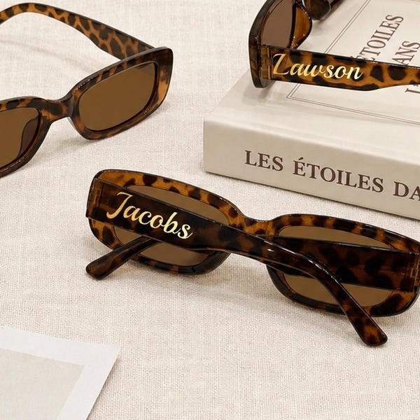 Op de afbeelding: Rechthoekige zonnebril met een bruin en zwart luipaardpatroon en donkerbruine glazen. Eén paar heeft de naam Jacobs in gouden schrijfletters op de poot, terwijl een ander paar de naam Lawson in gouden schrijfletters weergeeft.