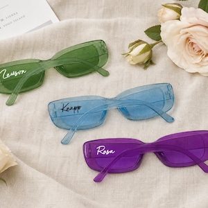 Peut inclure: Trois paires de lunettes de soleil rectangulaires en vert, bleu et violet translucides. La paire verte porte le texte en écriture blanche Lawson, la bleue le texte en écriture noire Knapp et la violette le texte en écriture blanche Rosa.