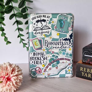May include: A clear e-reader case adorned with stickers. The stickers display phrases such as "You had me at dragons" and "Romantasy reader", alongside floral and fantasy-themed illustrations. The case has a blue and white checkered pattern.