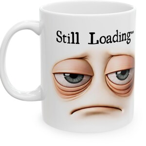 May include: White ceramic mug with a handle featuring a cartoon face with tired eyes and the text "Still Loading". The face has blue eyes, and the mug is suitable for hot or cold beverages.