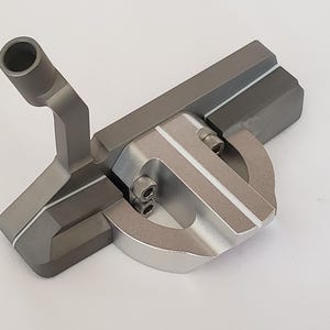 Könnte beinhalten: Ein grauer und silberner Golf-Putter mit rundem Kopf und rohrförmigem Schaft. Der Putter hat weiße Akzentlinien und sichtbare Schrauben. Das Design ist modern und elegant, geeignet für Golf-Enthusiasten.