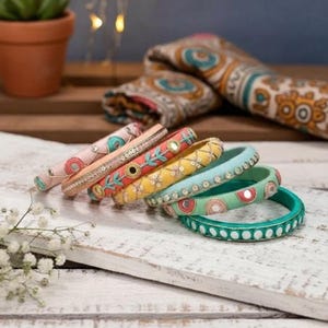Puede incluir: Una colección de pulseras coloridas con intrincados bordados y detalles de espejo. Las pulseras, en tonos rosa, amarillo, azul y verde, están dispuestas sobre una superficie de madera blanca. Una bufanda estampada es visible en el fondo.