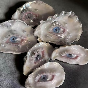 May include: Six oyster shells, each containing a realistic human eye sculpture. The eyes feature blue irises, pink eyelids, and fine eyelashes. The shells are pearlescent white with small, pearl-like details.