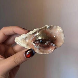 May include: An oyster shell decorated with a painted human eye featuring a metallic gold eyelid and a dark iris. The shell is embellished with several small, white, spherical beads of varying sizes scattered across the surface, creating a textured appearance.