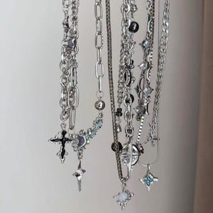 May include: A collection of silver-toned metal necklaces hanging vertically. The chains feature various link styles, including cable and box chains, adorned with charms such as black enamel crosses, iridescent stones, and star-shaped pendants.
