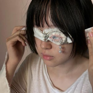 May include: A white fabric blindfold featuring a central realistic eye illustration, embellished with silver-toned metal hardware, small white beads, and a dangling strand of white pearls that hangs down from the lower edge of the eye opening.