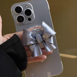 May include: A light gray smartphone with a triple-lens camera system on the back, featuring a prominent, three-dimensional, metallic silver bow-shaped phone grip attached to the center of the device's clear protective case.