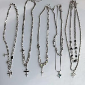 May include: Five silver-toned metal necklaces arranged side-by-side. Each necklace features a unique chain design, including link chains and delicate strands, adorned with various charms such as crosses, stars, and small beads in metallic tones.