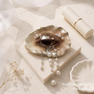May include: A decorative oyster shell featuring a painted human eye in the center, adorned with small white pearls and three dangling silver chains with pearl accents. The text Blue Tide Creation Shell oracle is printed in the bottom right corner.