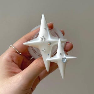 Peut inclure: Support de téléphone blanc en forme d'étoile avec des accents de strass. Le support présente deux étoiles de tailles différentes, chacune avec des pointes et des strass incrustés de différentes couleurs. Le support est conçu pour être fixé à l'arrière d'un téléphone.