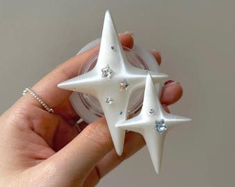 May include: White star-shaped phone grip with rhinestone accents. The grip features two stars of different sizes, each with pointed tips and embedded rhinestones in various colors. The grip is designed to attach to the back of a phone.