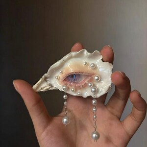 May include: An oyster shell featuring a realistic painted human eye with a blue iris and long eyelashes. The shell is decorated with multiple small white pearls embedded around the eye, and two silver chains with dangling pearls hang down.