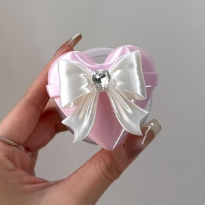 May include: A light pink heart-shaped object featuring a smooth, glossy finish. Centered on the front is a white satin ribbon bow, which is adorned with a single, clear, heart-shaped rhinestone at its center point, adding a reflective detail.