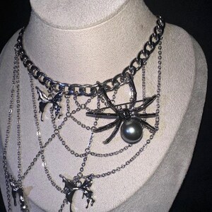 May include: Silver-toned necklace featuring a large spider pendant with a dark gray pearl, set within a web design. The necklace includes dangling chains and butterfly charms, displayed on a mannequin.