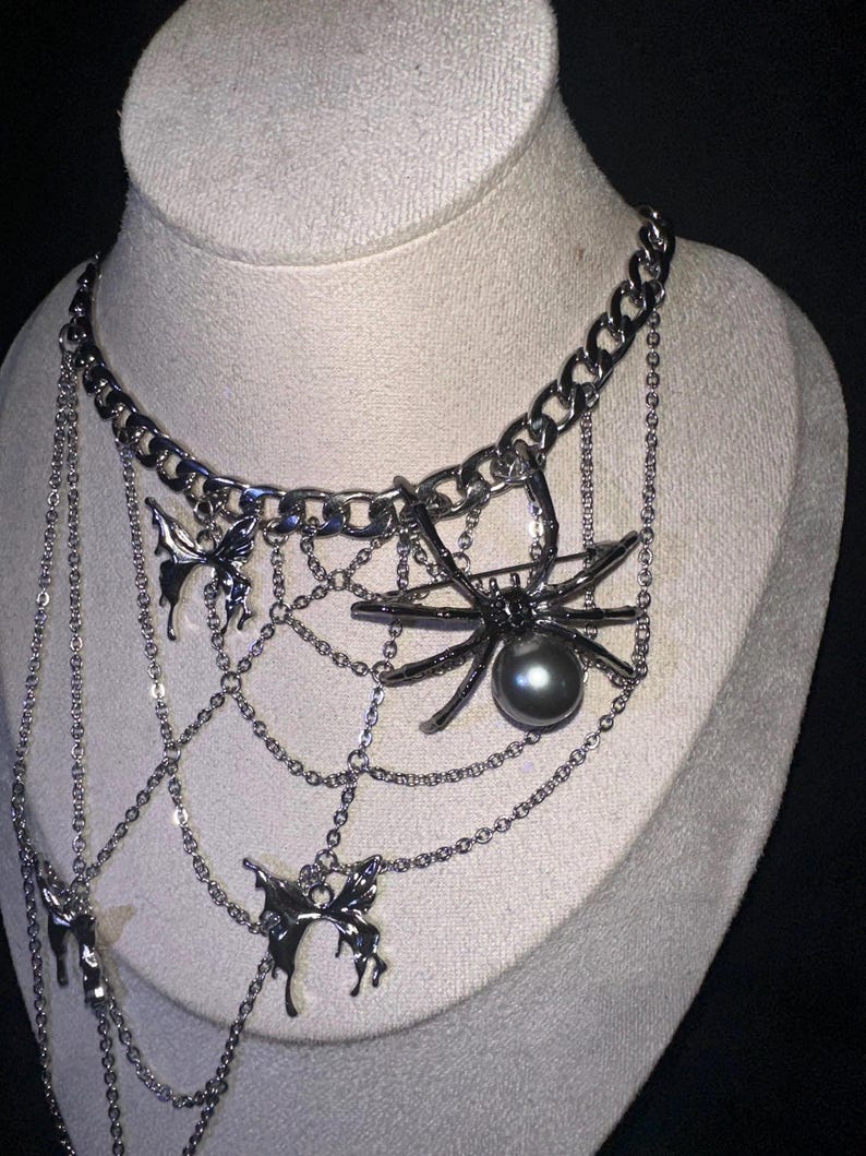 May include: Silver-toned necklace featuring a large spider pendant with a dark gray pearl, set within a web design. The necklace includes dangling chains and butterfly charms, displayed on a mannequin.
