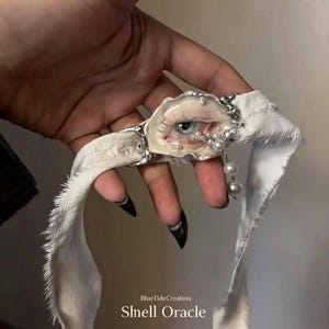Op de afbeelding: Een witte stoffen choker met een centraal schelp-ornament met een realistisch oogontwerp, geaccentueerd door kleine witte parels en zilverkleurige metalen details. De tekst BlueTideCreation Shell Oracle staat in het wit onderaan gedrukt.