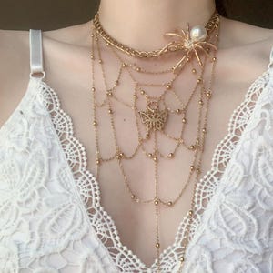 Könnte beinhalten: Goldfarbene Halskette mit einem Spinnenanhänger mit Perle, einem Schmetterlingsanhänger und einem spinnennetzartigen Design. Die Halskette hat mehrere Stränge aus zarten Ketten und Perlenakzenten, die einen geschichteten Effekt erzeugen. Der Schmuck wird über einem weißen Spitzentop getragen.