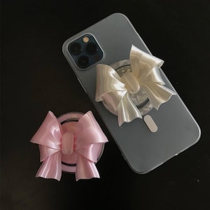 May include: A clear phone case with a pearlescent bow phone grip attached. A separate pink bow phone grip is also visible. The bows have a shiny, satin-like appearance. The phone case is on a dark surface.