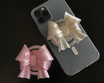May include: A clear phone case with a pearl-colored bow phone grip attached. A separate pink bow phone grip is also visible. The bows have a shiny, satin-like appearance. The phone case is on a dark surface.